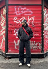 Homme en coupe-vent noir et pantalon, portant un bonnet et des lunettes de soleil, debout devant un rideau métallique rouge couvert de graffitis.