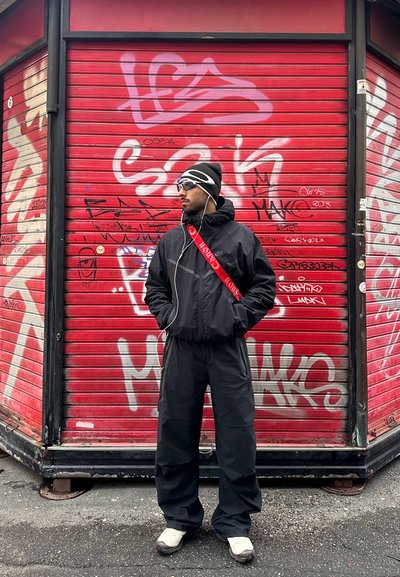 Hombre con cortavientos negro y pantalones, con gorro y gafas de sol, de pie frente a una persiana metálica cubierta de graffiti rojo.