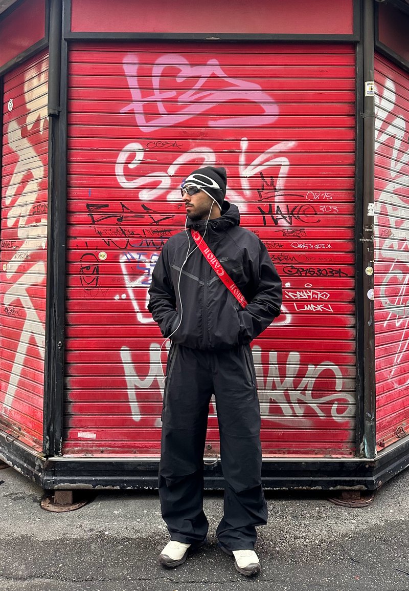 Homme en coupe-vent noir et pantalon, portant un bonnet et des lunettes de soleil, debout devant un rideau métallique rouge couvert de graffitis.