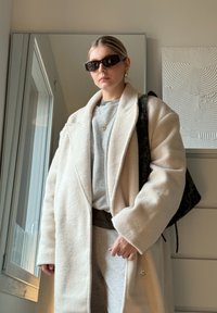 Casaco oversized creme texturizado, t-shirt cinza, calças de moletom cinzas, óculos de sol pretos e uma mala com padrão preto, contra um fundo neutro.