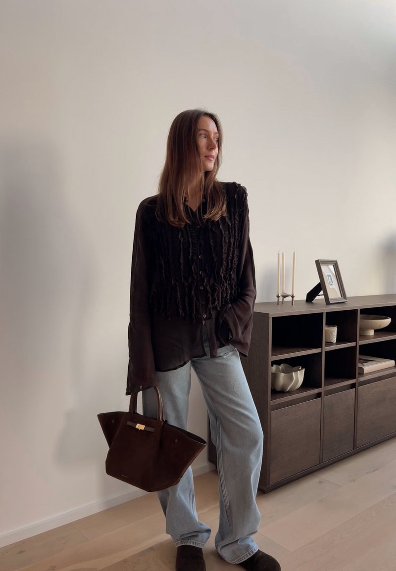 Camisa preta com pregas e botões, jeans de perna larga azul-claro e uma bolsa de camurça castanho-escuro. Unidade de armazenamento em madeira ao fundo.