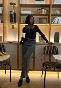 Maglietta nera a maniche lunghe, pantaloni grigi a gamba larga, sciarpa grigia, in mano una tazza di caffè bianca, in piedi accanto a uno scaffale in legno con oggetti decorativi.