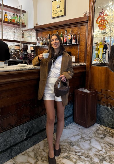 Blazer de ante marrón sobre una blusa blanca y pantalones cortos, sosteniendo una taza de café. Bolso trenzado marrón. Ambiente de bar con acentos de madera y estanterías.