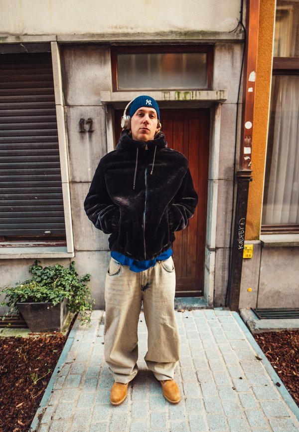 Giacca nera in pile con cappuccio e zip, camicia blu sotto, pantaloni beige larghi e stivali color cuoio. Cuffie bianche e berretto blu indossati.