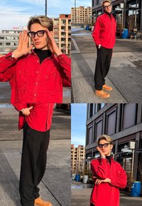 Jeune personne aux cheveux blonds courts et portant des lunettes noires pose en plein air, habillée d'une chemise rouge, d'un pantalon noir et de bottes beige, près de bâtiments urbains.