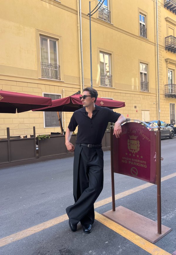 Černé polo tričko s krátkým rukávem, široké černé kalhoty a lesklé černé boty. Opírá se o červenou značku "Žádné parkování". Jasné městské prostředí.
