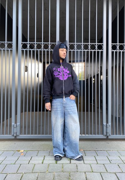 Sudadera con cremallera negra y gráfico morado, combinada con jeans de pierna ancha de color azul desvanecido y sandalias negras, de pie frente a una puerta de metal.