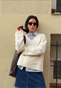 Une femme portant des lunettes de soleil, un pull crème, une chemise rayée et une jupe en jean se tient contre un mur beige, tenant un sac marron et jouant avec ses cheveux.