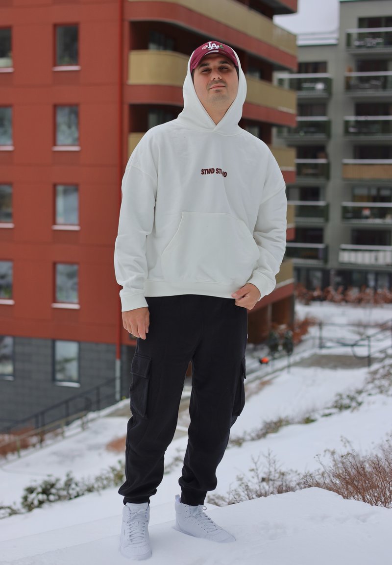 Jeune homme portant un sweat à capuche blanc, un pantalon cargo noir, des baskets blanches et une casquette bordeaux, debout à l'extérieur dans une zone urbaine enneigée.