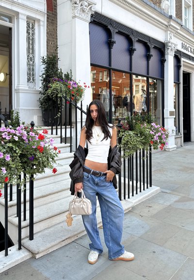 Top corto blanco, chaqueta bomber marrón oversized, jeans azules holgados y zapatillas blancas. Sosteniendo un pequeño bolso crema con un charm.