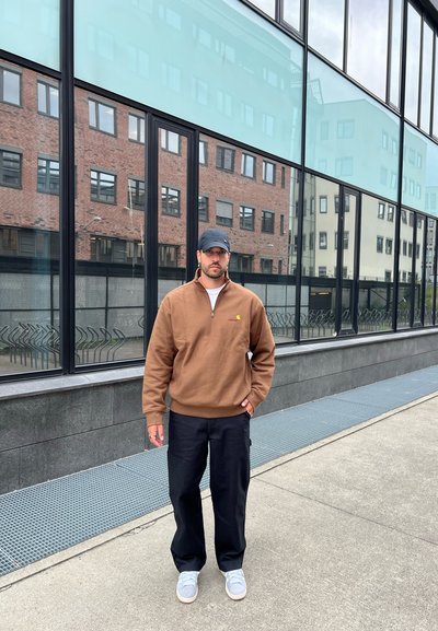 Sudadera marrón con cremallera y un pequeño logo, pantalones cargo negros y zapatillas grises. Tejido suave y corte relajado, de pie al aire libre.