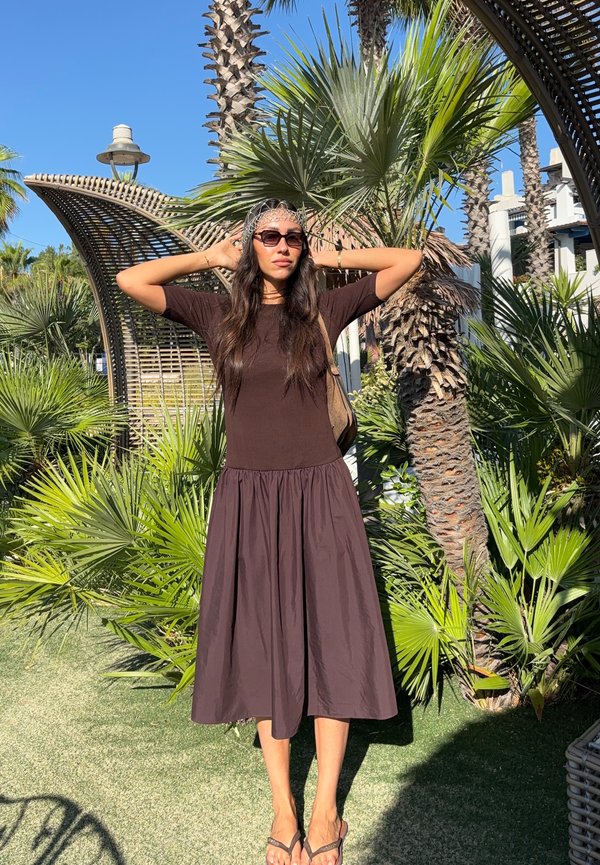 Femme portant des lunettes de soleil et une robe sombre se tient les mains derrière la tête parmi des palmiers sous un ciel bleu clair.