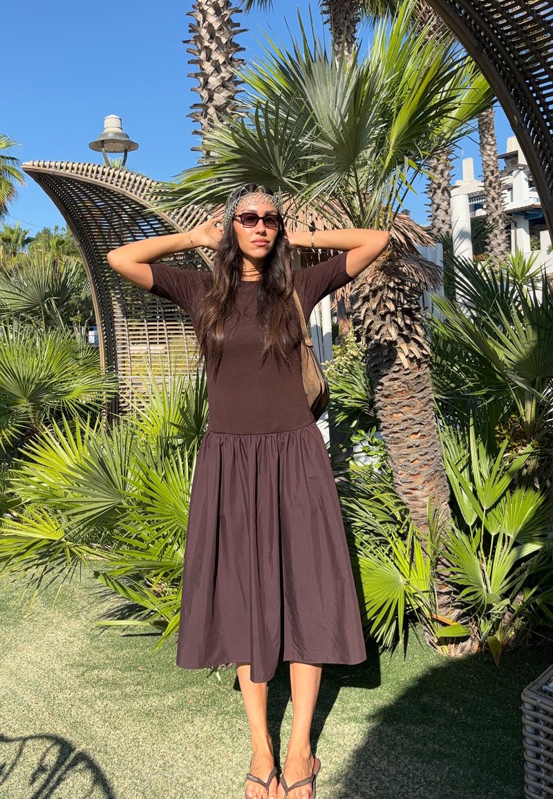 Femme portant des lunettes de soleil et une robe sombre se tient les mains derrière la tête parmi des palmiers sous un ciel bleu clair.