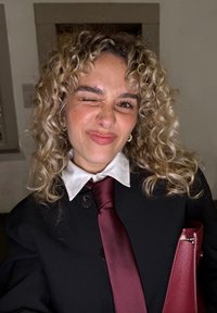 Czarna marynarka z dużymi guzikami, biała koszula z kołnierzykiem i bordowy krawat z satyny. Kręcone włosy; trzyma bordową skórzaną kopertówkę.