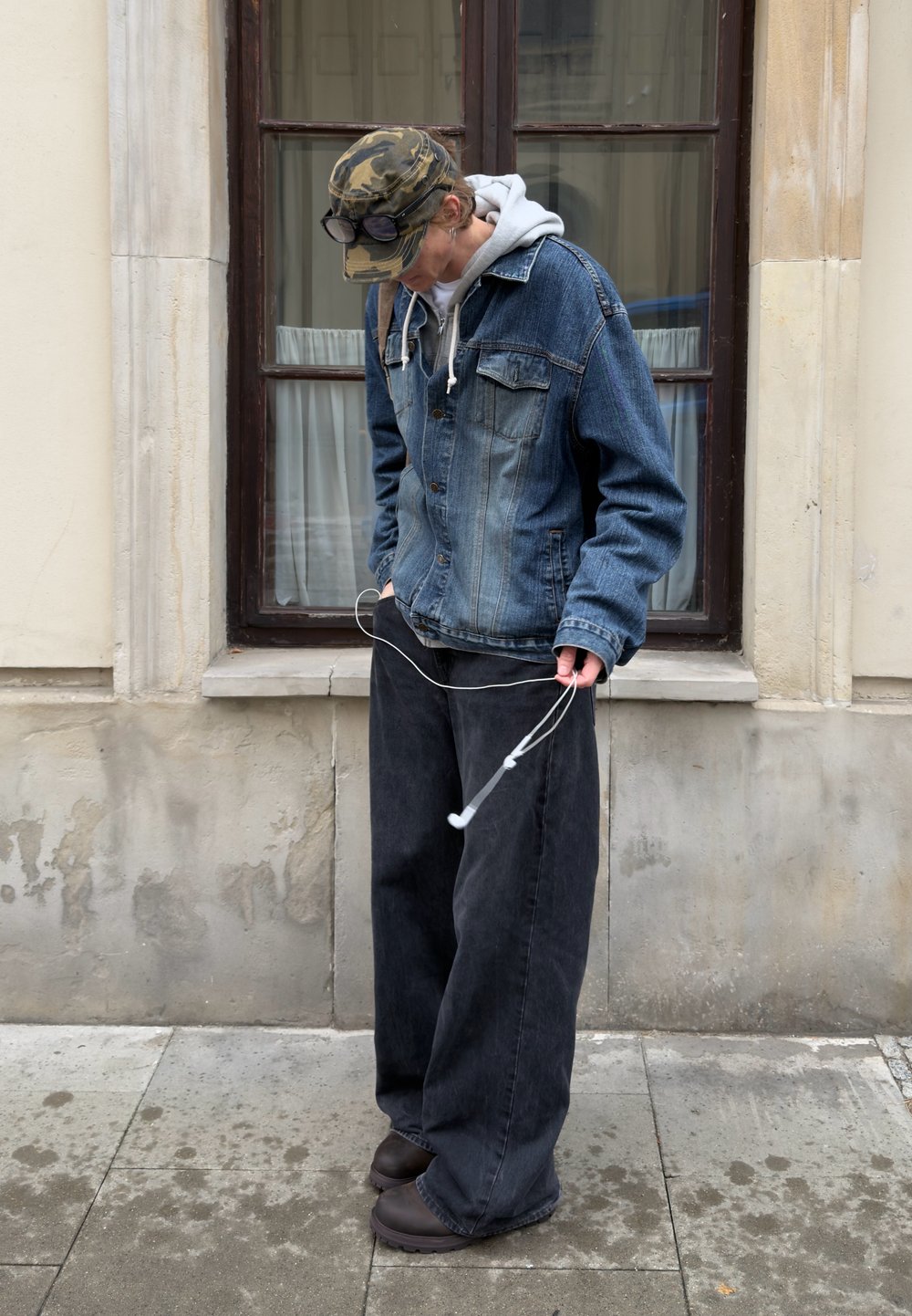 Jeansjacka med två bröstfickor, grå hoodie under, lösa svarta jeans och mörka skor. Vita hörlurar hålls i ena handen.