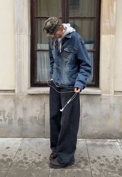 Chaqueta de denim con dos bolsillos en el pecho, sudadera gris debajo, jeans negros anchos y zapatos oscuros. Auriculares blancos sostenidos en una mano.