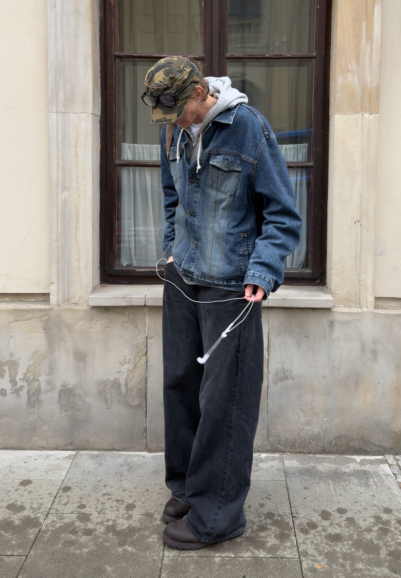 Denimjakke med to brystlommer, grå hættetrøje under, baggy sorte jeans og mørke sko. Hvite høretelefoner holdt i den ene hånd.