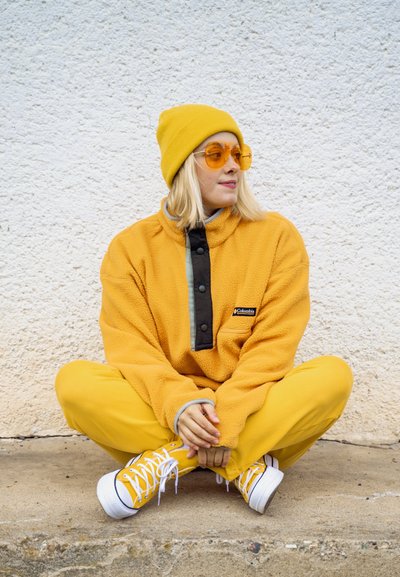 Sudadera de felpa amarilla con tapeta de botones negros y acentos grises, combinada con pantalones amarillos y zapatillas amarillas. Gafas de sol con tinte naranja y gorro.