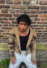 Brązowa kurtka z baranią wełną z beżowym wnętrzem, noszona na czarnej koszuli i białych spodniach. Siedzi oparty o ceglany mur z zielenią w tle.