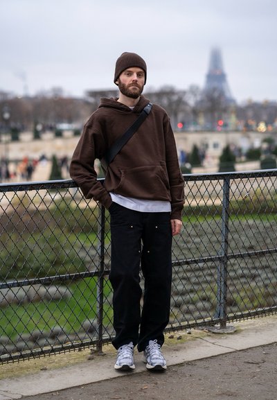 Sudadera con capucha marrón, pantalones oscuros con bolsillos, zapatillas de deporte grises. Gorro marrón, de pie junto a una valla con un fondo desenfocado de vegetación.