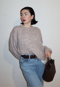 Femme aux cheveux courts et foncés portant un pull beige et un jean bleu, tenant un sac à main tressé foncé, regardant vers sa gauche contre un mur uni.