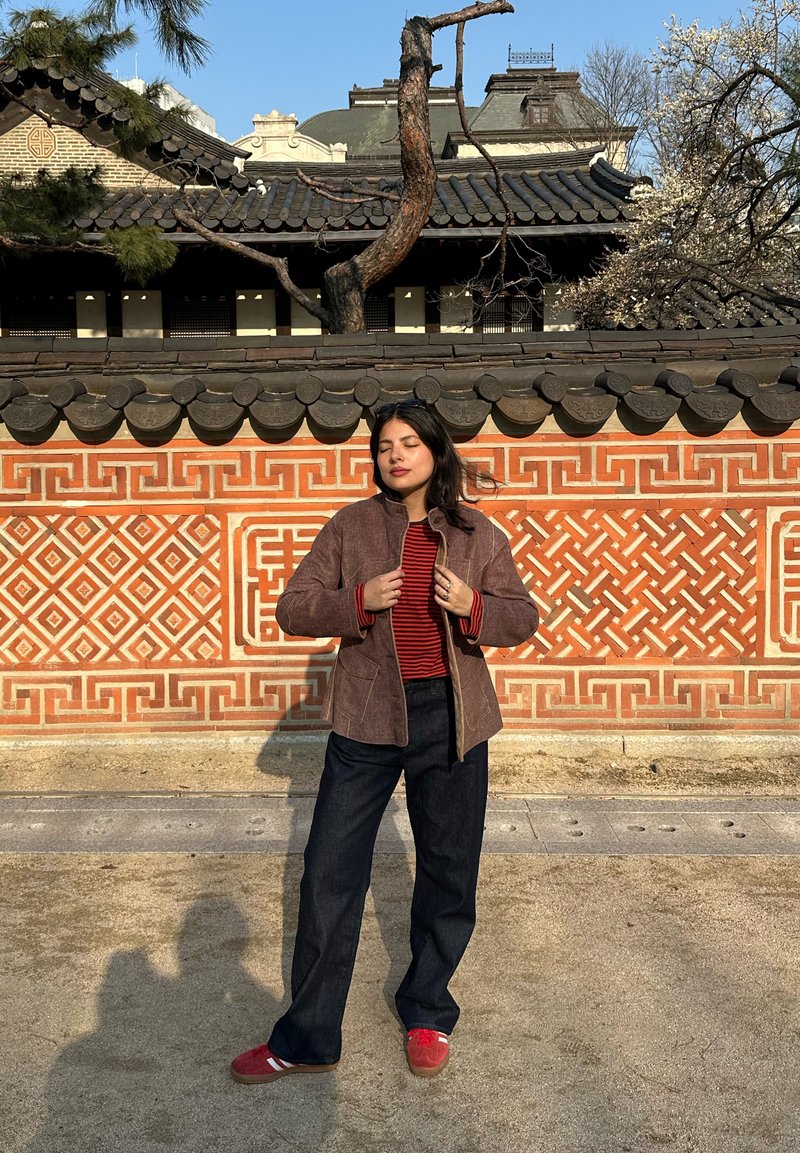 Žena stojící se zavřenýma očima před dekorativní stěnou s geometrickým vzorem a tradiční korejskou střechou na slunečním světle.