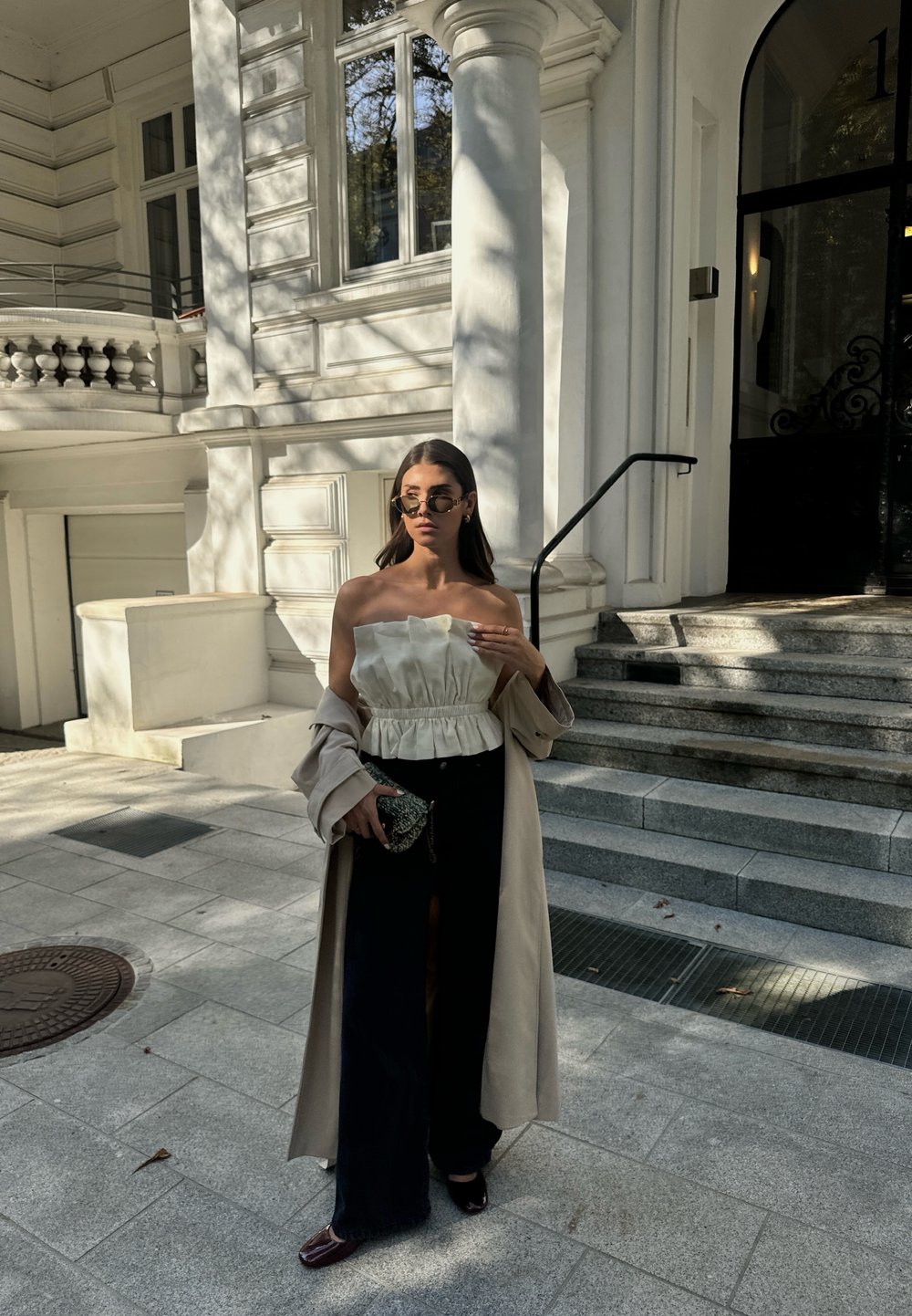 Μπεζ παλτό σε στυλ trench πάνω από λευκή τοπ χωρίς τιράντες με υφή και ρίγες, συνδυασμένο με μαύρο παντελόνι φαρδύ και σκούρα παπούτσια από πατινέ δέρμα.