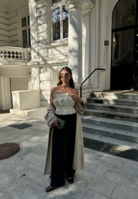 Μπεζ παλτό σε στυλ trench πάνω από λευκή τοπ χωρίς τιράντες με υφή και ρίγες, συνδυασμένο με μαύρο παντελόνι φαρδύ και σκούρα παπούτσια από πατινέ δέρμα.