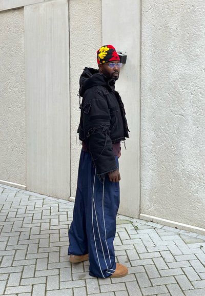 Chaqueta negra corta con bordes deshilachados, combinada con pantalones holgados azul marino con rayas blancas y calzado marrón. Lleva puesta una gorra de punto con un estampado colorido.