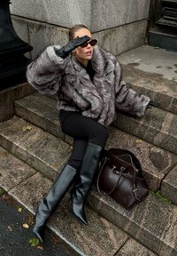 Szary płaszcz z futerka sztucznego, dopasowane czarne spodnie, czarne kozaki do kolan oraz ciemna torebka. Siedzi na kamiennych schodach, nosi okulary przeciwsłoneczne i rękawiczki.