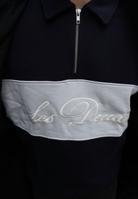 Ναυτικό μπλε φούτερ με φερμουάρ μέχρι τη μέση, λευκή οριζόντια λωρίδα και ανεβασμένο λευκό κεντητό κείμενο "Les Deux" στο στήθος.