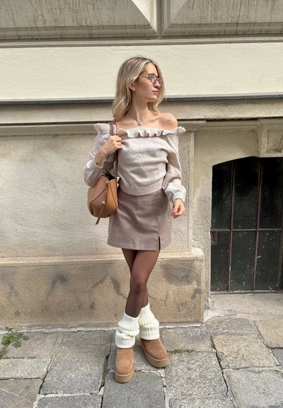 Suéter crema con escote rizado, falda corta de cuadros, botas de tobillo color beige y calentadores de piernas crema. Bolso marrón con correa ajustable.