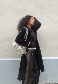 Mulher com cabelo encaracolado vestindo um casaco longo preto, calças listradas em preto e branco e a carregar uma mala de ombro branca diante de uma parede pálida.