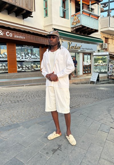 Una persona está de pie en una calle de adoquines, vistiendo una camisa blanca de manga larga, pantalones cortos de cargo beige y zuecos de un color claro.