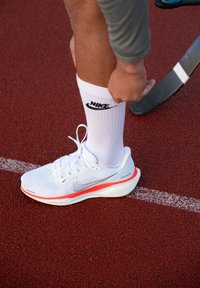 Hvit sportssko med sølvfarget swoosh og oransje såle, kombinert med en hvit sokk med svart Nike-logo, på en rød løpebane.