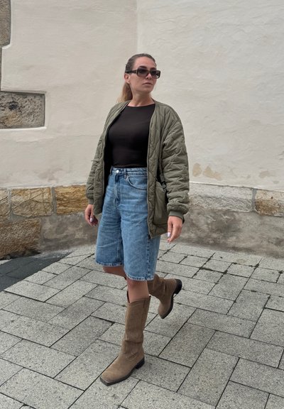 Chaqueta bomber acolchada de color verde oliva, blusa negra de manga larga, pantalones cortos de bermuda de mezclilla azul claro y botas vaqueras marrones hasta la rodilla.