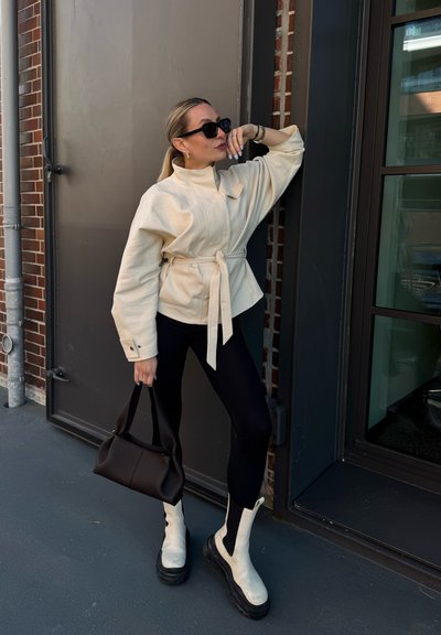 Mujer con chaqueta beige con cinturón, leggings negros, botas blancas y gafas de sol, sosteniendo un bolso negro, posando frente a una pared de ladrillo con puerta de cristal.