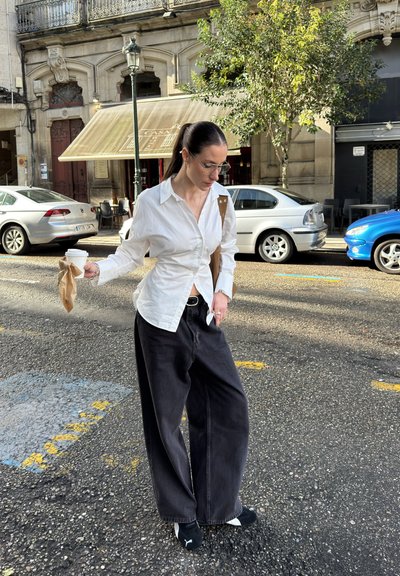 Camisa blanca de corte clásico, pantalones anchos negros, sosteniendo una taza de café en papel marrón. Zapatos negros de diseño deportivo. Entorno urbano.