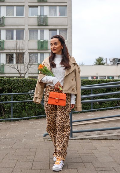 Abrigo trench beige, suéter blanco de punto, pantalones de leopardo, bolso naranja y zapatillas blancas, sosteniendo tulipanes, en un entorno urbano al aire libre.