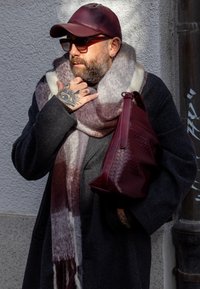 Viininpunainen nahkalaukku, harmaa huivi valkoisilla ja viininpunaisilla sävyillä, harmaa päällystakki, tummat aurinkolasit ja viininpunainen lippalakki. Kädessä näkyvät tatuoinnit.