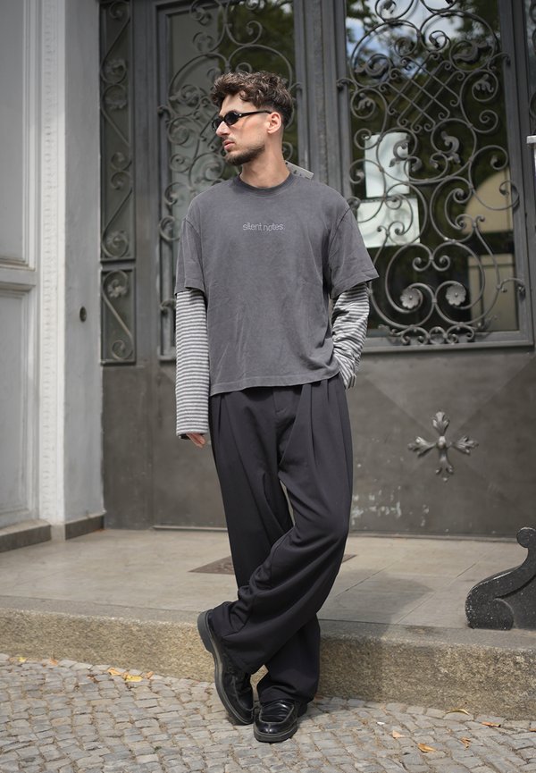 Homme portant des lunettes de soleil foncées, des manches longues rayées grises superposées et un t-shirt, pantalon noir, debout jambes croisées près d'une porte en métal ornée.