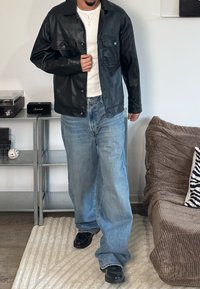 Mustast nahast jakk kahe rinna taskuga, kantud valge t-särgi kohal. Lahtised sinised teksad ja mustad jalanõud. Neutraalse ilmega eluruumi taust.