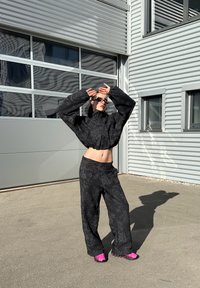 Jaqueta cropped com padrão floral preto, com mangas oversized, combinada com calças largas. Modelo usa calçado rosa-choque, posando ao ar livre.