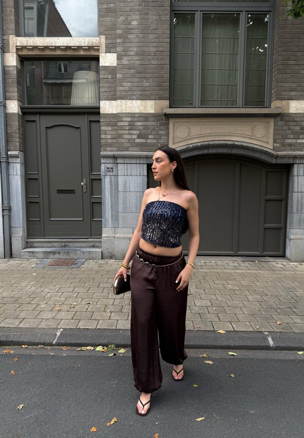 Femme marchant dans une rue devant un bâtiment en briques, portant un haut noir sans bretelles, un pantalon large marron et tenant une petite pochette.