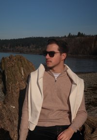 Νεαρός άντρας που φοράει γυαλιά ηλίου και μπεζ πουλόβερ με λευκό μπουφάν περασμένο στους ώμους κάθεται πάνω σε μεγάλο κορμό ξύλου δίπλα σε λίμνη με δάσος στο βάθος.