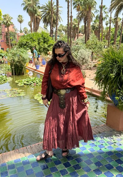 Blusa de lino roja suelta, falda burdeos acampanada y fluida con detalles metálicos. De pie junto a un estanque rodeado de palmeras y vegetación.