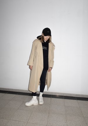 Mujer con gabardina beige, sudadera con capucha negra, leggings negros, zapatillas blancas y gorra negra, de pie sobre suelo de baldosas frente a una pared blanca.