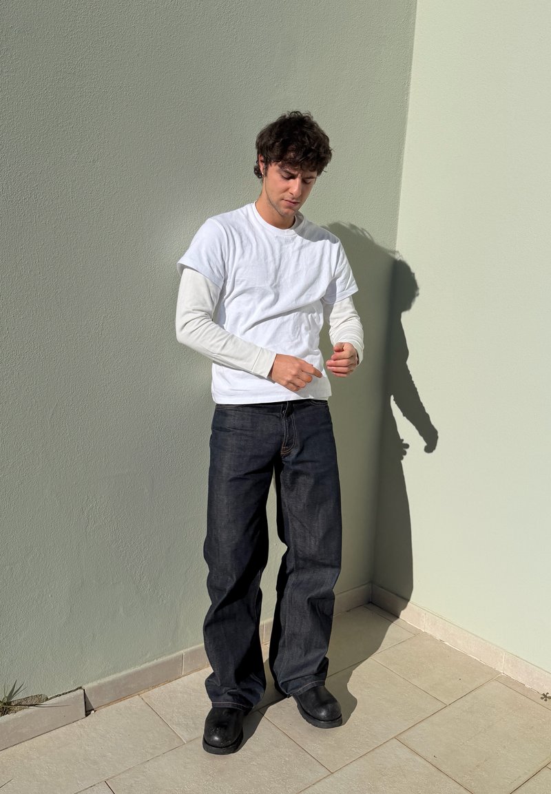 Ung mann som står inntil et hjørne, iført en hvit langermet skjorte under en hvit kortermet skjorte, mørke jeans og svarte sko.