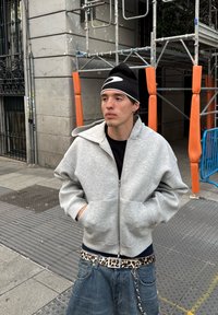 Pelēka kapucēta jakas ar lielu apmali, valkāta virs melnas krekla. Zili džinsi ar leopardu apdruku jostu. Melns beanie cepure ar baltu logotipu.