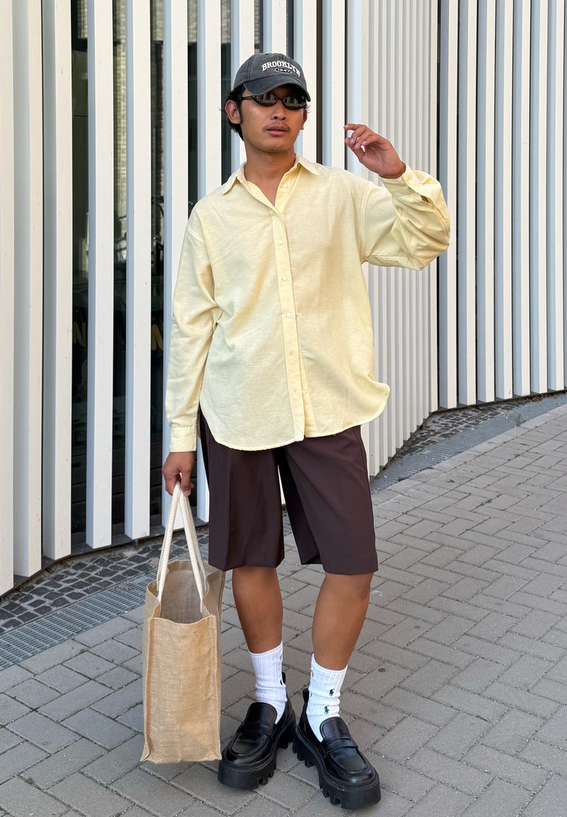 Helles gelbes Langarmhemd mit Knöpfen, braune Shorts, schwarze Plateauschuhe, weiße Socken und eine beigefarbene Tragetasche, dazu Sonnenbrille und Kappe.